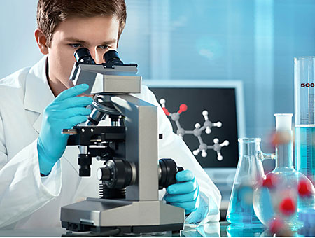 Een onderzoeker in een witte labojas zit achter een moderne, binoculaire microscoop. Hij kijkt door de lenzen. Hij draagt blauwe nitril handschoenen. Verder op de voorgrond zien we glazen flessen een reageerbuis. In de achtergrond een scherm met daarop de chemische structuur van de één of andere stof.